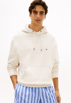 Junger Mann trägt einen cremefarbenen Hoodie mit kleinem Logo, kombiniert mit blau-weiß gestreifter Hose, steht mit den Händen in den Taschen.