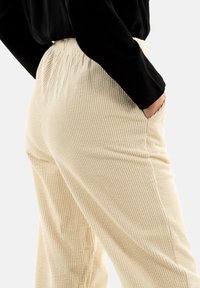 Beige corduroy broek met verticale ribbels, elastische tailleband en zijzakken, gepresenteerd tegen een neutrale achtergrond.