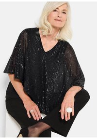 Schwarzer Pailletten-Poncho mit V-Ausschnitt und weiten Ärmeln, über einem schwarzen Oberteil und einer Hose getragen. Hat eine glatte Textur und schimmernde Akzente.
