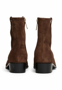 Stivaletti alla caviglia in suede marrone con una superficie texturizzata, tacco in gomma nera e dettagli in zip dorata sul retro. Design semplice e moderno.