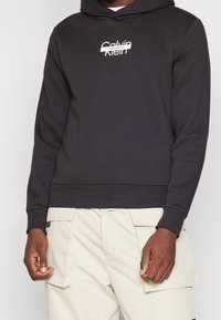 Felpa nera con cappuccio realizzata in materiale morbido, con un logo bianco di Calvin Klein sul petto, polsini a costine e una vestibilità rilassata.