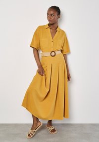 Gelbes, kurzärmliges Kleid mit Knopfverschluss und zwei vorderen Taschen, an der Taille mit einem gewebten Gürtel mit einer runden Schnalle gerafft.