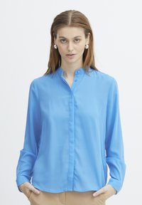 Camicia a maniche lunghe azzurro chiaro con colletto a camicia, bottone frontale, texture liscia e vestibilità comoda. Le mani del modello sono nelle tasche, design minimale.