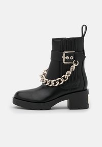 Stivaletto in pelle nera con una suola chunky, con un accento di catena metallica e una fascia con fibbia. Design a righe verticali sul lato.