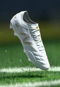 Pólo de futebol branco com um upper texturizado, apresentando um logótipo e atacadores em destaque dourado. Ramalhetes de relva estão deslocados sob o sapato.