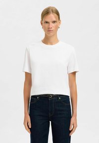 Valkoinen puuvillapaita lyhyillä hihoilla, pyöreällä kaula-aukolla ja rentoa mittatilausta vastaavalla leikkauksella, yhdistetty tummansinisiin farkkuihin ja mustaan vyöhön. Sileä rakenne.