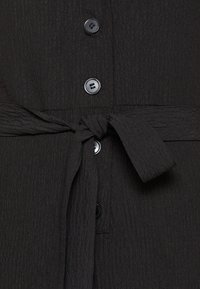 Detailní záběr na tmavý strukturovaný oděv se čtyřmi černými knoflíky a zavázaným látkovým páskem v oblasti pasu.