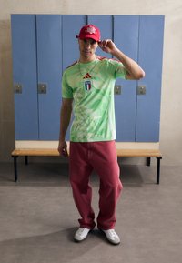 Žalias grafiškas T-shirt su lapų raštais ir mėlynu Italijos ženkleliu, derinamas su laisvomis raudonomis kelnėmis ir baltomis sportinėmis bateliais, pristatomas prieš spintas.