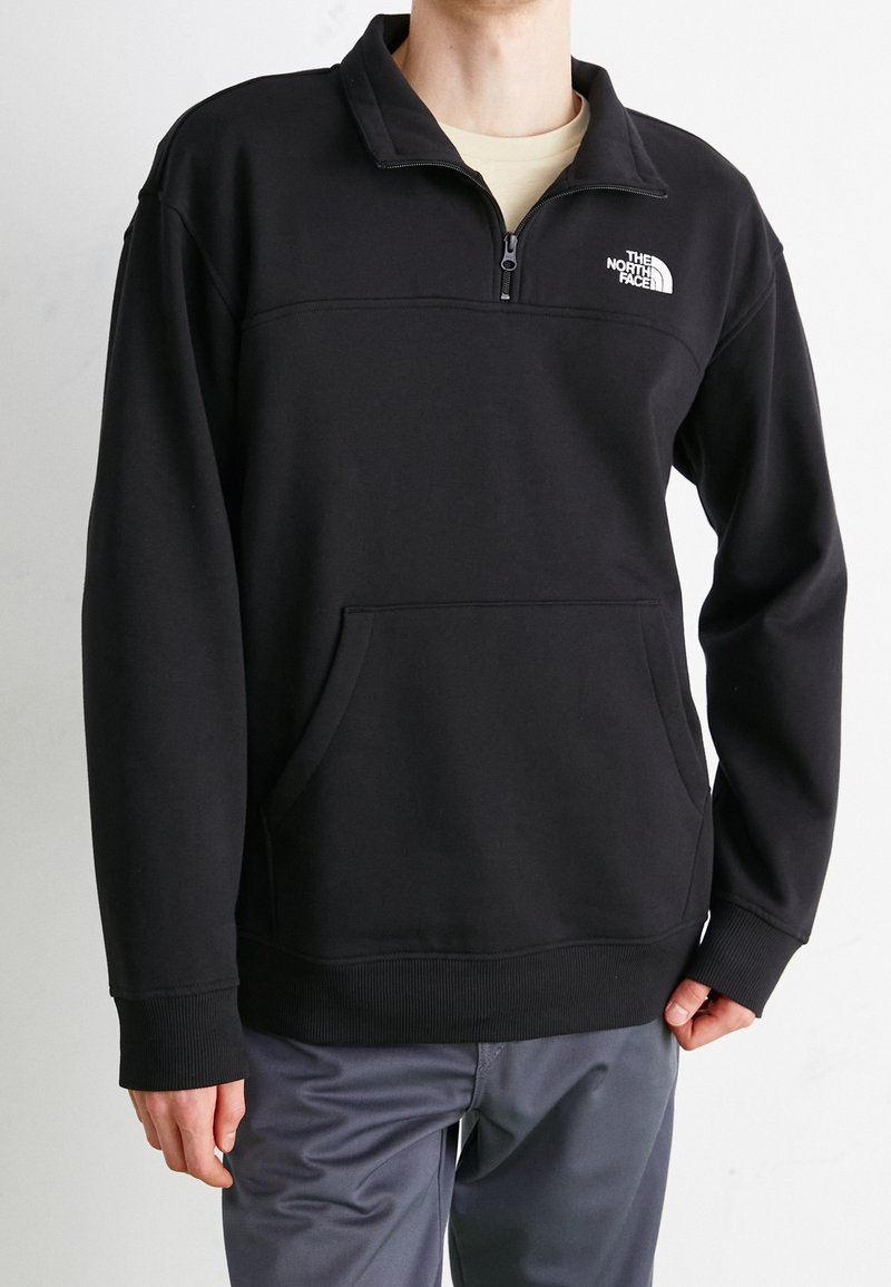 Pullover in pile nero con colletto a zip a un quarto, tasca frontale e polsini e orlo a costine. Presenta un logo bianco sul petto.