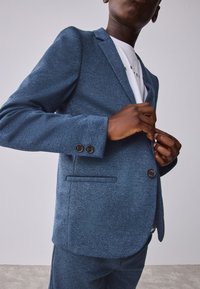 Blazer azul oscuro con textura y patrón de espiga; presenta solapas con muesca, dos botones y bolsillos laterales. Llevado sobre una camisa blanca.