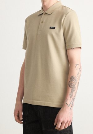 Mann trägt beiges kurzärmeliges Polohemd mit kleinem schwarzem Calvin Klein-Label auf der Brust und sichtbarem Schlangentattoo am rechten Unterarm.