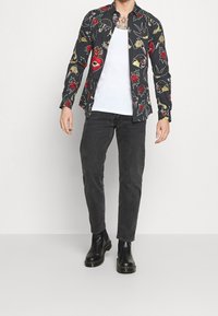 Chemise noire à boutons ornée d'imprimés floraux et graphiques colorés. Portée sur un débardeur blanc, associée à un jean skinny noir et des bottes noires.
