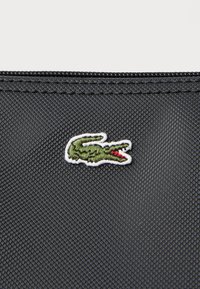 Un tissu noir texturé présente un petit logo crocodile vert brodé avec des détails rouges. Les coutures sont visibles le long des bords.