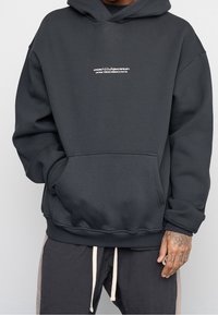 Ciemnoszara, oversize bluza z kapturem wykonana z miękkiego materiału, z kieszenią kangurzą z przodu i małym białym graficznym logo na klatce piersiowej.