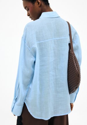 Mujer de espaldas, vestida con una camisa azul clara de gran tamaño y llevando un bolso de piel marrón tejido en el hombro derecho.