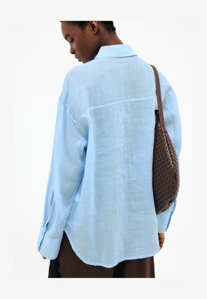 Žena otočená chrbtom, oblečená v nadmernej svetlomodrej košeli, nesie hnedú pletenú koženú kabelku na pravom ramene.