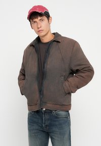 Brown padded jacket with a zip front, featuring a collar and side pockets. Worn over a dark shirt, paired with blue faded jeans and a pink cap.