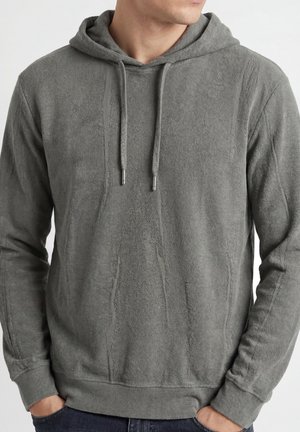 Hombre con una sudadera con capucha gris texturizada con cordones, manos en los bolsillos, mirando ligeramente hacia la derecha.