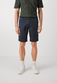 Shorts blu navy in cotone, con tasche laterali, taglio dritto, orli piegati e chiusura con bottone indossati da un modello con una camicia verde.