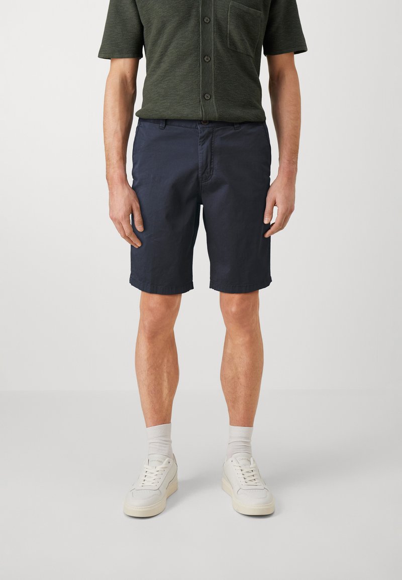 Shorts blu navy in cotone, con tasche laterali, taglio dritto, orli piegati e chiusura con bottone indossati da un modello con una camicia verde.