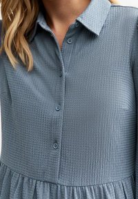 Blå knäppt skjorta med en texturerad tyg, med en krage och en samlad nederkant. Tre knappar synliga på framsidan.