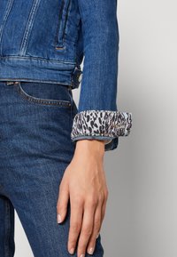 Chaqueta de denim con un lavado azul, con puños doblados que revelan un forro de estampado animal en gris claro. Jeans ajustados complementan el conjunto.