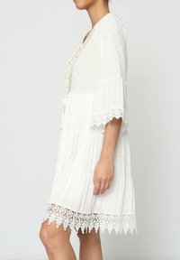 Robe blanche à manches courtes, au design à étages, avec un ourlet en dentelle. Présente des motifs dorés et un lien tressé au niveau du col.