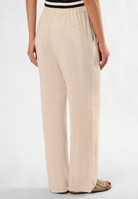 Beige weite Hose aus strukturiertem Stoff mit elastischem Bund und Gesäßtaschen. Kombiniert mit hellbraunen Sandalen.