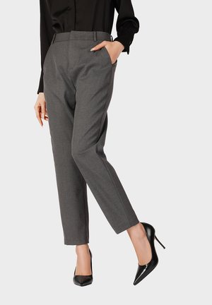 Mujer con pantalones grises a medida y zapatos negros de tacón alto, mano en el bolsillo, sobre un fondo liso.