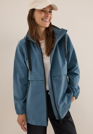 Blauwe lichte jas met een ritssluiting aan de voorkant, zakken en elastische manchetten. Gepaard met een lichtgekleurde top en een beige pet. Gladde textuur.