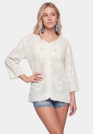 Femme blonde portant un chemisier blanc transparent brodé de fleurs avec manches trois quarts et boucles d'oreilles à pompons bleues, associé à un short en jean.