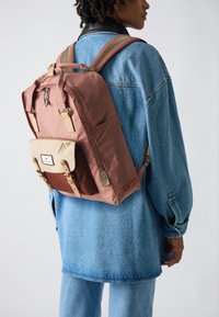 Sac à dos rose, marron et crème, en matériau en tissu, avec des bretelles ajustables, une poche avant avec rabat et des accents en matériel, porté sur une tenue en denim.