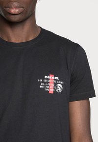 Svart bomull t-shirt med tryckt logotyp i röd och vit text, rund halsringning och korta ärmar. Texten inkluderar "DIESEL" och produktinformation.
