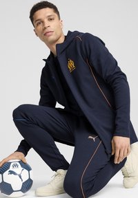 Giacca a vento blu navale con dettagli arancioni e logo, abbinata a pantaloni coordinati. Tiene un pallone da calcio ed indossa sneakers color crema.