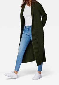 Lång, öppen front cardigan i mörkgrön ribbad stickning, bärs över en vit topp och blå skinny jeans, i kombination med vita sneakers.