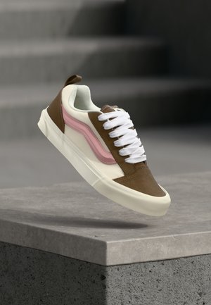 Brun, hvid og pink sneaker med hvide snørebånd svævende over en grå stenkant, med slørede grå trin i baggrunden.