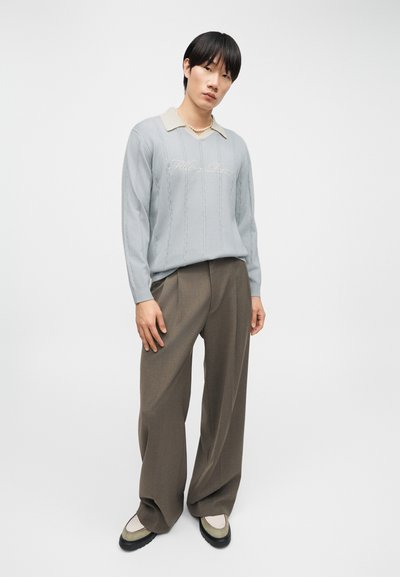 Νεαρός άνδρας που φοράει ανοιχτό μπλε πλεκτό πουλόβερ με κείμενο, μπεζ πουκάμισο με γιακά, φαρδιές καφέ παντελόνες, κολιέ με πέρλες και δίχρωμα παπούτσια, όρθιος.