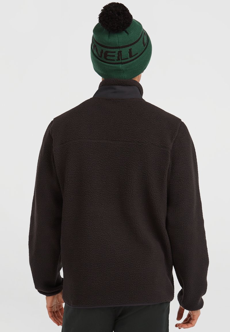 Uomo che indossa una giacca in pile nera e un cappello di maglia verde con pompon nero, visto da dietro su uno sfondo chiaro e uniforme.