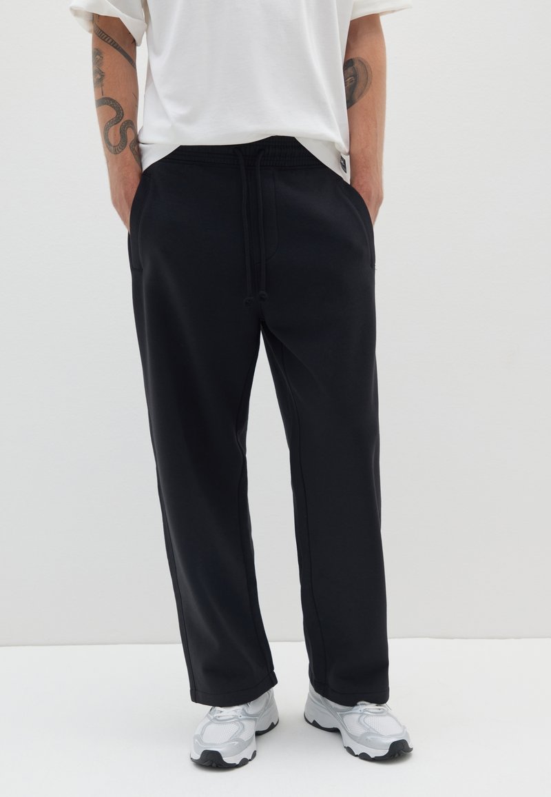 Schwarze Jogginghose mit elastischem Bund, Zugkordel, seitlichen Taschen und geradem Schnitt. Kombiniert mit einem weißen T-Shirt und grauen Sportschuhen.