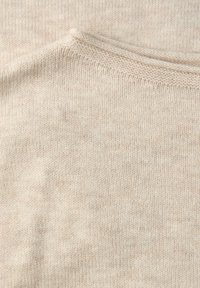 Beżowa dzianina o miękkiej fakturze, z delikatnym melanżowym wzorem. Widoczne przeszycia wzdłuż krawędzi i kołnierza.