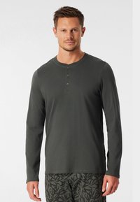 Maglietta henley a maniche lunghe in cotone misto verde scuro con colletto tondo e patta con tre bottoni, abbinata a pantaloni corti scuri con fantasia.