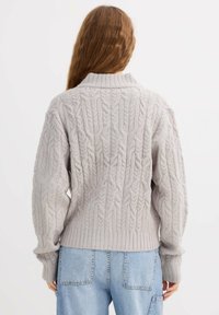 Suéter gris claro de punto trenzado con cuello alto y dobladillo acanalado, con patrones texturizados. Combinado con jeans azul claro de pierna ancha.