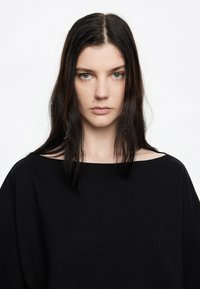 Czarna, oversize bluzka z dekoltem w łódkę i opadającymi ramionami, wykonana z gładkiego materiału. Modelka ma długie ciemne włosy i neutralny makijaż.
