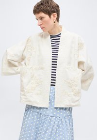 Femme aux cheveux courts portant des boucles d'oreilles dorées, une veste blanche brodée de fleurs par-dessus un haut rayé bleu marine et blanc, et une jupe bleue à pois.