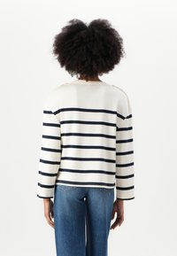 Stripet langermet topp i krem med marineblå horisontale striper, med skulderknapper og avslappet passform. Kombinert med denimjeans.