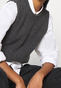 Suéter gris de punto sin mangas con textura acanalada, llevado sobre una camisa blanca con cuello. La persona está sentada con las manos reposando sobre las rodillas.