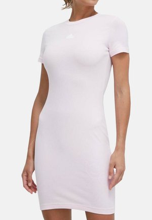Donna che indossa un abito aderente rosa pallido a maniche corte con un piccolo logo bianco sul petto, in piedi davanti a uno sfondo semplice.