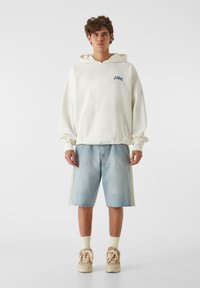 Gai bežs kapuce ar priekšējo kabatu un zilu logo, kombinēta ar gaiši ziliem džinsiem, kas ir īsi un ar sapīlētu maliņu un teksturētām sānu daļām.