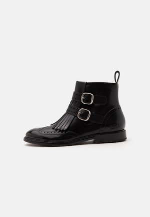 Bottine noire en cuir avec design wingtip, franges décoratives, deux boucles argentées et une languette à l'arrière, présentée sur fond blanc.