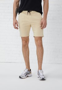 Beige shorts med snøre, der har en glat tekstur og opkrøllede manchetter, parret med sølvfarvede træningssko og en sort T-shirt.